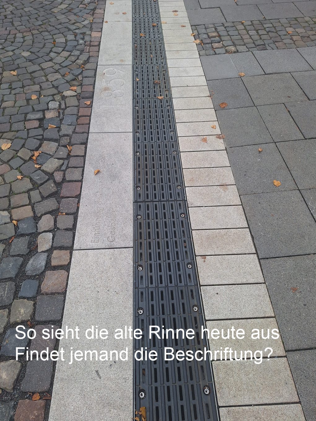 Die bisherige Regenablaufrinne mit dunklem Metallgitter, flankiert von hellen Steinplatten mit historischer Beschriftung, daneben Kopfsteinpflaster.