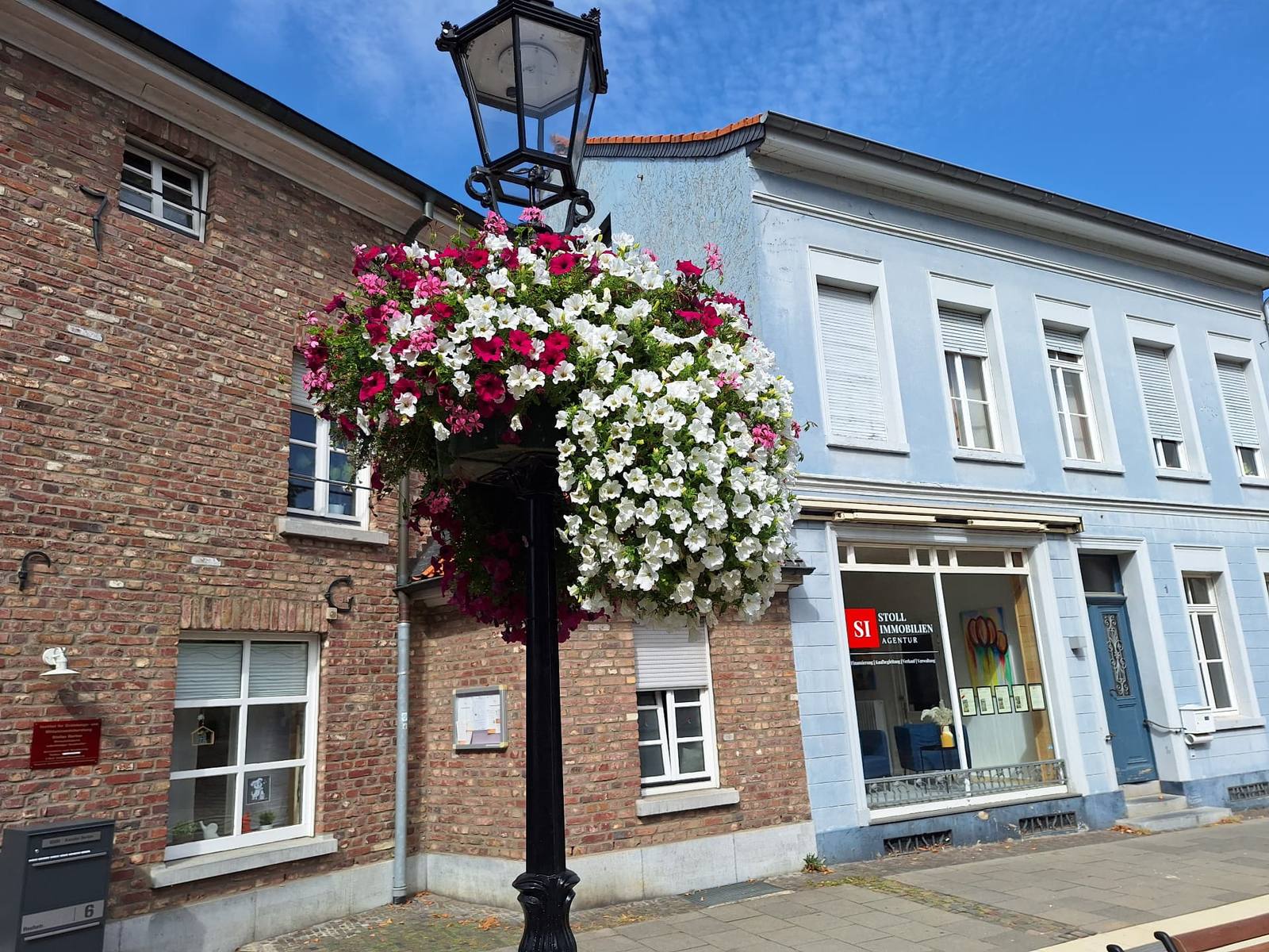 Schwarze Laternenstange mit üppigen Blumenampeln in Weiß und Pink vor historischen Häusern in Süchteln